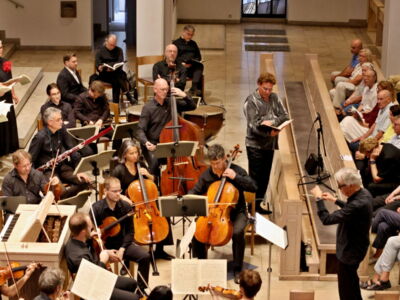 An orchestra and choir perform in a spacious hall, with musicians playing string instruments and a conductor leading. The audience watches attentively from pews on the right.