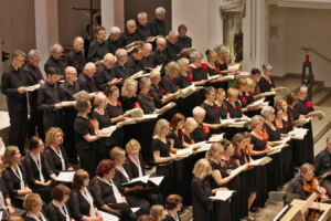 A large choir dressed in black with red accents performs in a tiered formation inside a church.