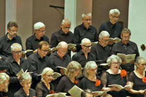 A choir of older adults dressed in black with orange accents sings together, holding sheet music.