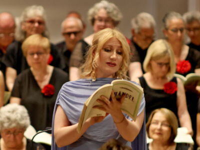 A woman sings from a music book in front of a choir, all dressed in black with red rose accessories.