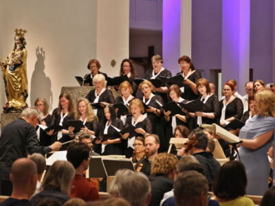 A choir performs in a church, with a conductor leading them and a soloist standing nearby. The audience is seated, and soft purple lighting illuminates the background.