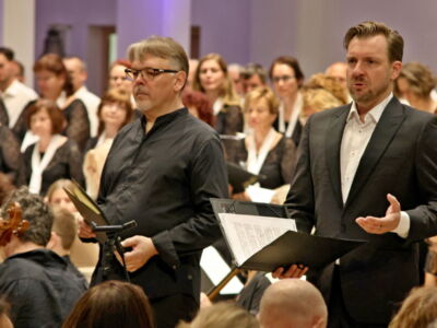 Two men in formal attire perform in front of a choir inside a concert hall.
