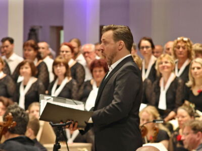 A man in a suit stands in front of a choir, holding a book. The choir members, dressed in black with white scarves, are focused on a performance or rehearsal in a brightly lit room.