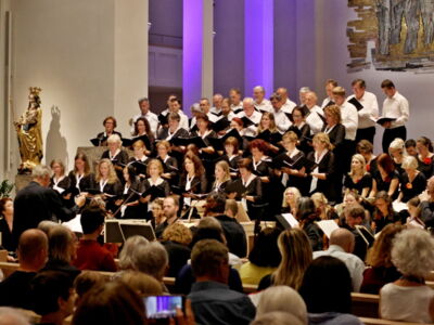 A choir performs in a well-lit church, with singers dressed in black and white. An audience watches from pews, and purple lighting enhances the setting.