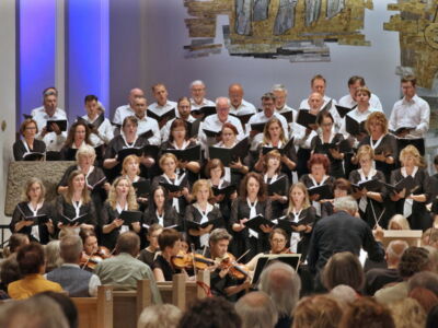 A choir performs in a church setting, with singers dressed in black and white, accompanied by musicians. A conductor stands in front, facing the group. The audience is seated, watching the performance.