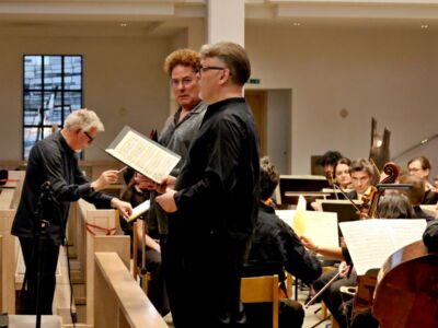 Musicians and singers rehearse with sheet music in a concert hall, surrounded by an orchestra.