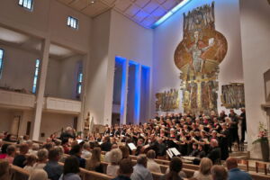 A large choir performs in a spacious church with a high ceiling and ornate wall art. The audience sits attentively, facing the choir. Soft lighting enhances the serene atmosphere.