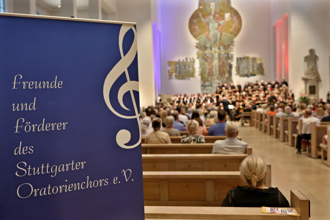 A choir concert taking place in a church, with people seated and a blue banner displaying text in German in the foreground.