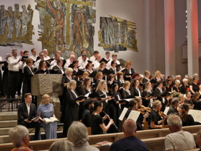A choir and orchestra perform in a church with an audience watching. The choir stands on risers, and musicians are seated below. Wall art decorates the background.