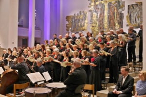 A choir performs in a grand hall, surrounded by musicians with instruments. The singers, holding songbooks, stand in front of an ornate backdrop with artistic wall reliefs.