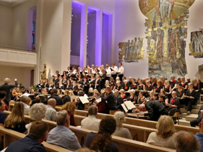 A large choir performs onstage in a well-lit church with a colorful mural. An attentive audience sits in wooden pews, watching the conductor in the foreground.