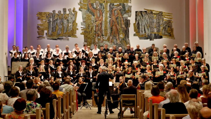 A choir performs in a church, with a conductor leading them. The audience watches from wooden pews, and the backdrop features artistic wall decorations.