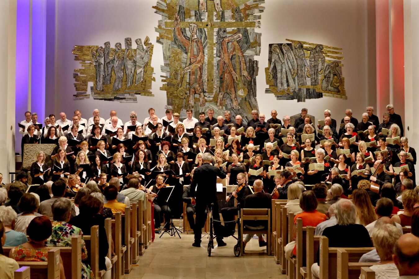 A choir performs in a church, with a conductor leading them. The audience watches from wooden pews, and the backdrop features artistic wall decorations.